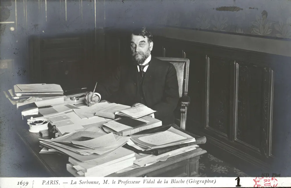 Le professeur Vidal de la Blache assis à son bureau de la Sorbonne posant le stylo à la main. Le bureau est couvert de livres, d’atlas, de brouillons d’articles, avec, bien en vue, un carnet de terrain